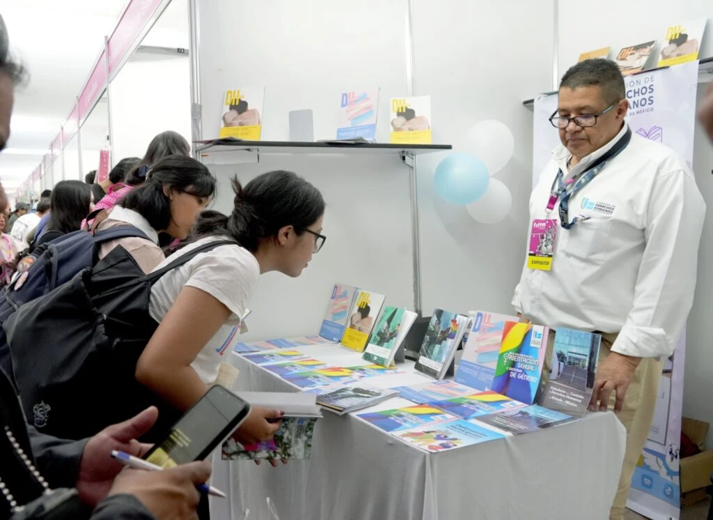 Varias personas consultan publicaciones y materiales informativos en un stand de la Comisión de Derechos Humanos del Estado de México. En la mesa se exhiben folletos, revistas y guías temáticas relacionadas con derechos humanos.