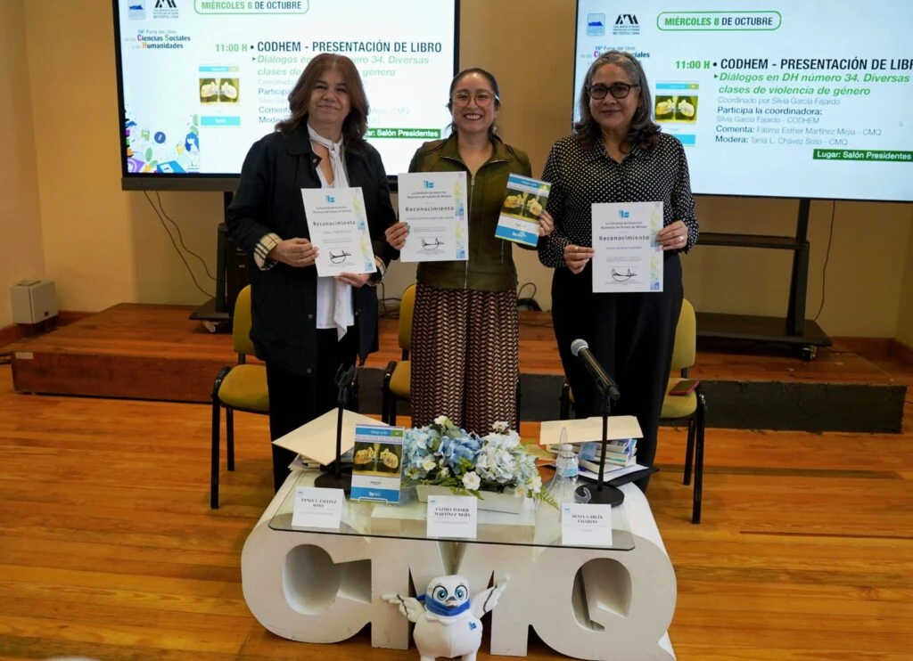 Tres personas posan de pie frente a dos pantallas de proyección durante una presentación de libro. Cada persona sostiene reconocimientos impresos y una de ellas también muestra un ejemplar del libro. En las pantallas se aprecia información del evento relacionado con la presentación del libro “Diálogos en DH número 34. Diversas clases de violencia de género”.