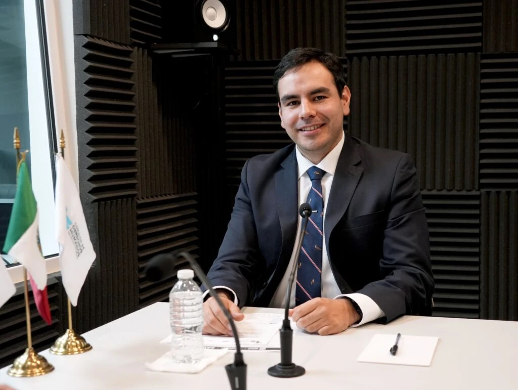 Persona participante en una entrevista o grabación institucional dentro de una cabina acústica, sentada frente a micrófonos y documentación, con banderas institucionales visibles al fondo.