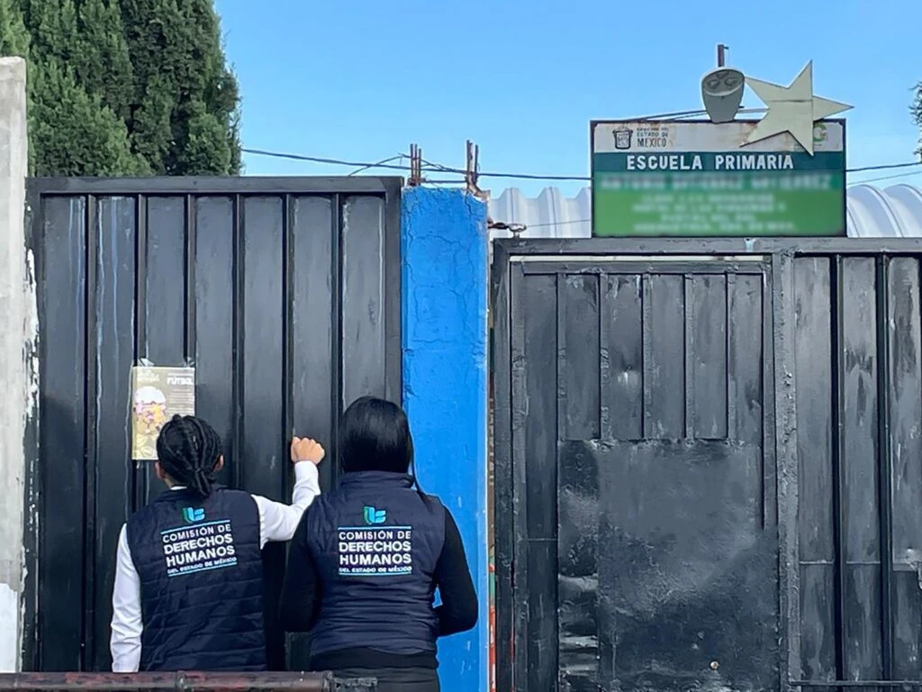 Dos personas de espaldas frente a la entrada de una escuela primaria con portón metálico negro y una columna azul. Ambas llevan chalecos azul marino con el texto “Comisión de Derechos Humanos del Estado de México”. En la parte superior derecha aparece un letrero verde de “Escuela Primaria” bajo un cielo despejado.