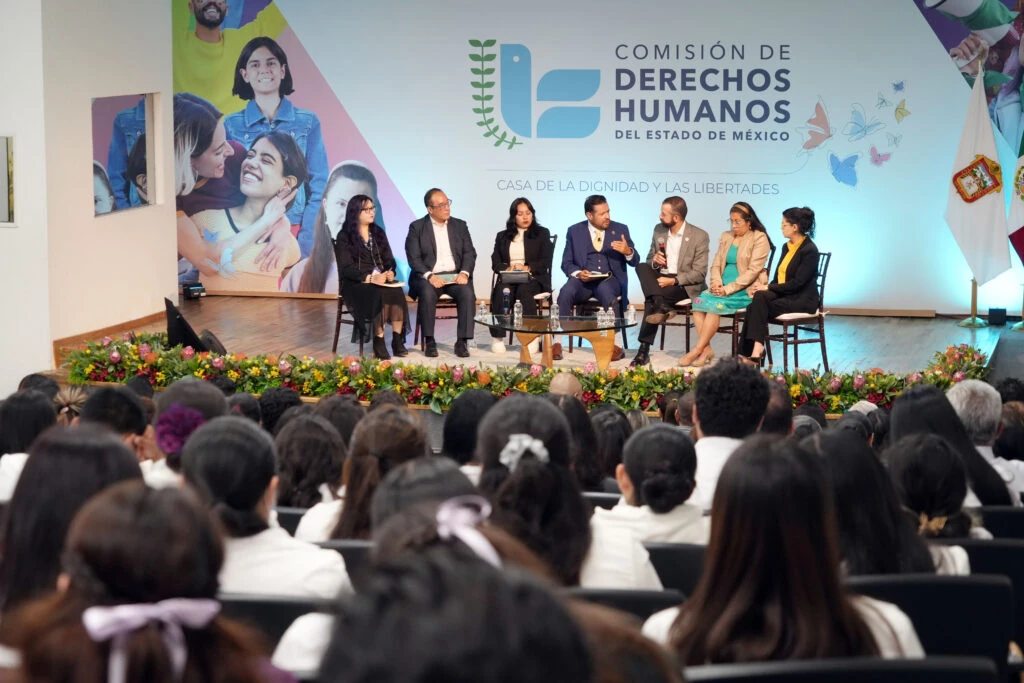 Vista amplia de un panel en el escenario de un auditorio, frente a un público numeroso. Varias personas están sentadas en sillas alrededor de una mesa central con botellas de agua y micrófonos. El escenario está decorado con arreglos florales y al fondo aparece el logotipo de la Comisión de Derechos Humanos del Estado de México junto al texto “Casa de la Dignidad y las Libertades”.