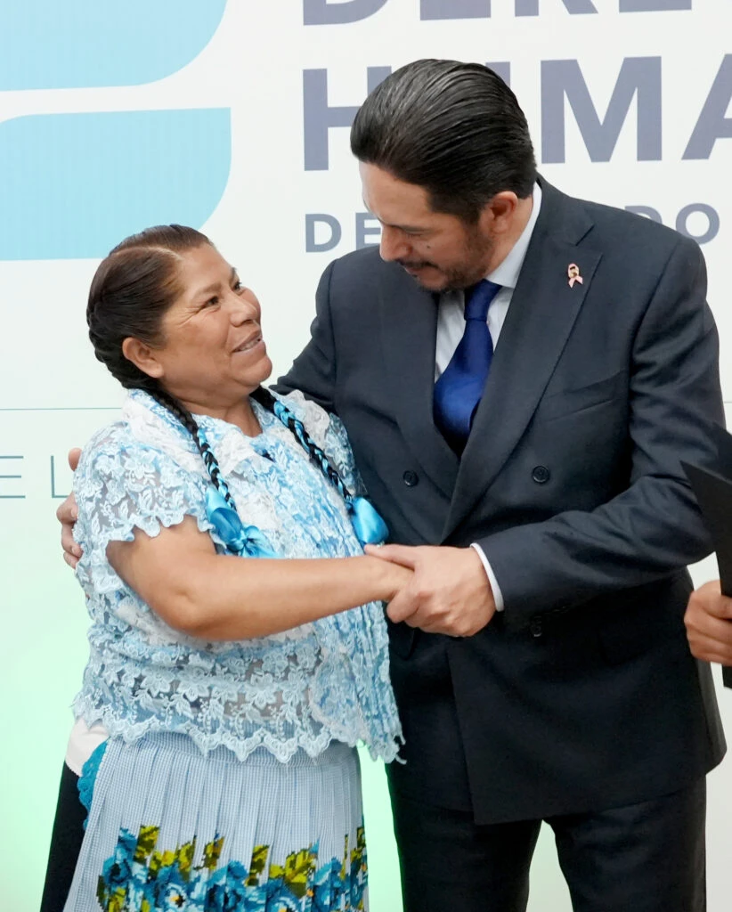 El presidente de la CODHEM Victor Leopoldo Delgado Pérez, estrecha la mano de otra persona que viste una blusa azul con detalles florales y falda tradicional. Ambas se encuentran frente a un fondo institucional con parte del logotipo y el texto de la Comisión de Derechos Humanos del Estado de México visibles al fondo.