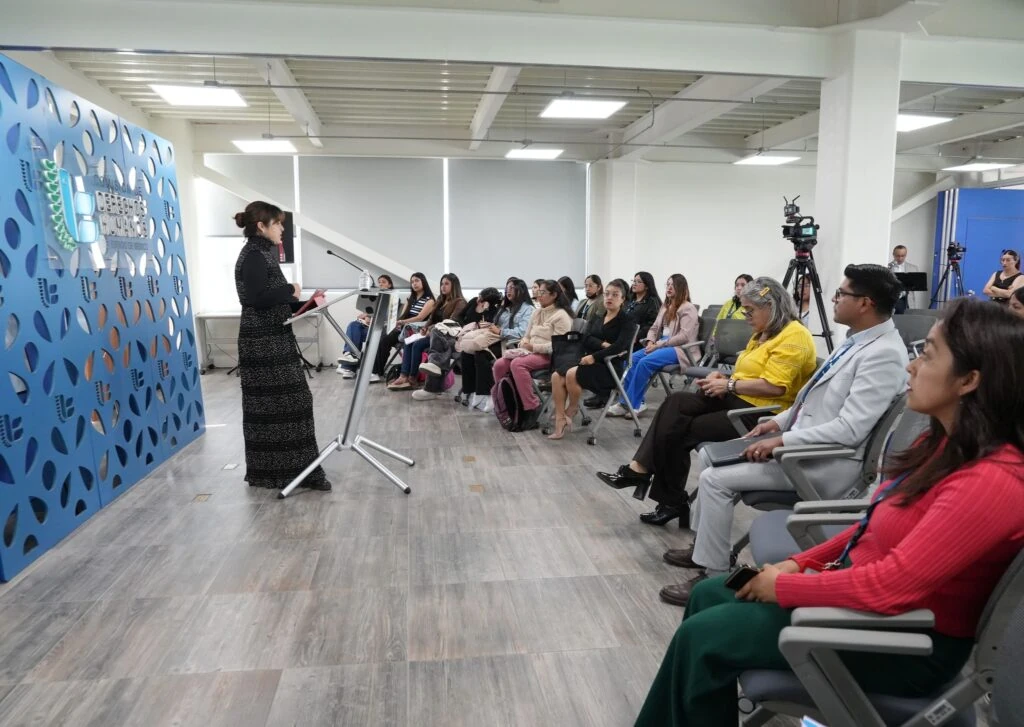 Vista amplia de una sala de conferencias con una persona de pie ante un atril y varias personas sentadas en filas. A la izquierda se observa un panel azul con el texto “Comisión de Derechos Humanos del Estado de México”, y al fondo hay cámaras en tripié y equipo de audio.
