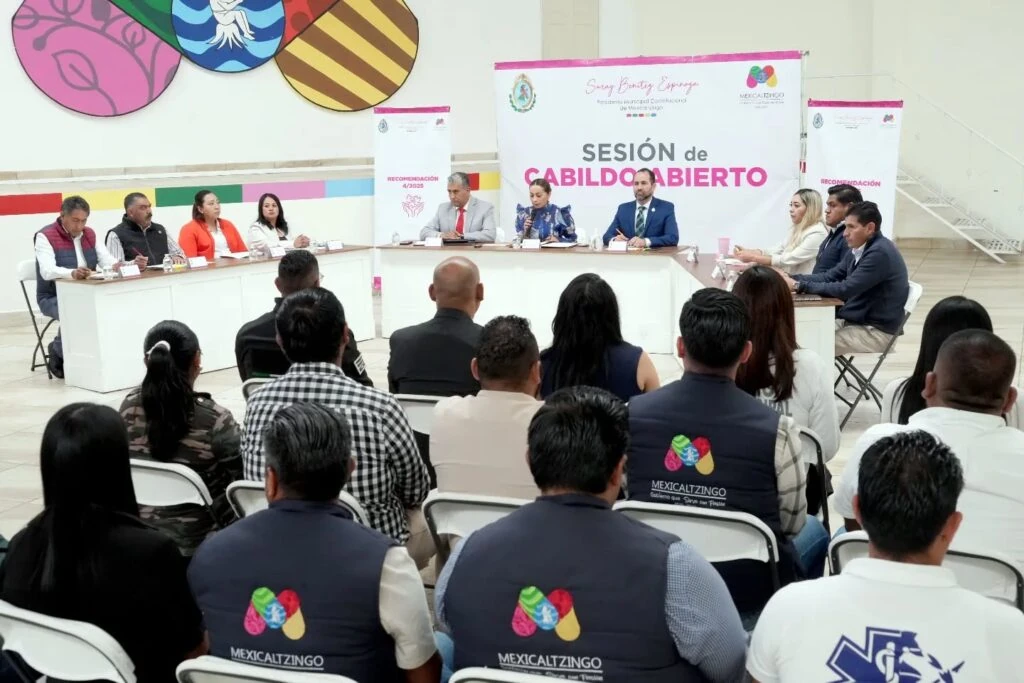Vista general de una sesión de cabildo abierto en un salón, con personas sentadas en mesas al frente y público en sillas blancas. Al fondo se observa una lona con el texto “Sesión de Cabildo Abierto” y elementos gráficos multicolores; varias personas del público portan chalecos con la palabra “Mexicaltzingo”.