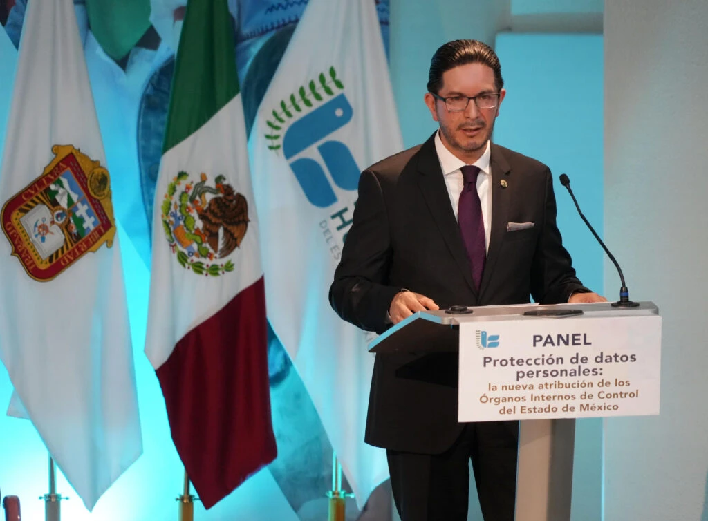 Protección de datos personales, acto de libertad y de respeto a la dignidad: Víctor Delgado Pérez. Una persona expositora interviene desde el atril durante el panel “Protección de datos personales: la nueva atribución de los Órganos Internos de Control del Estado de México”, con banderas institucionales al fondo.