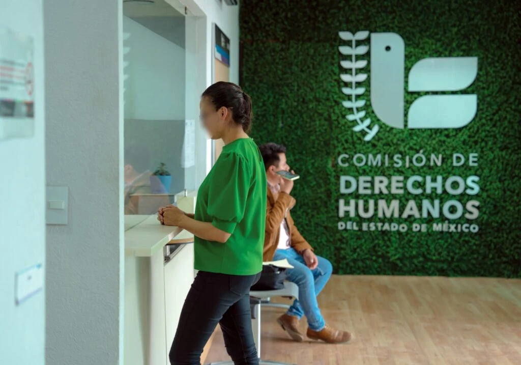 Texto alternativo: Persona revisando documentos en un módulo de atención de la Comisión de Derechos Humanos del Estado de México; detrás se aprecian paneles decorativos con el logotipo institucional y una planta de interior.