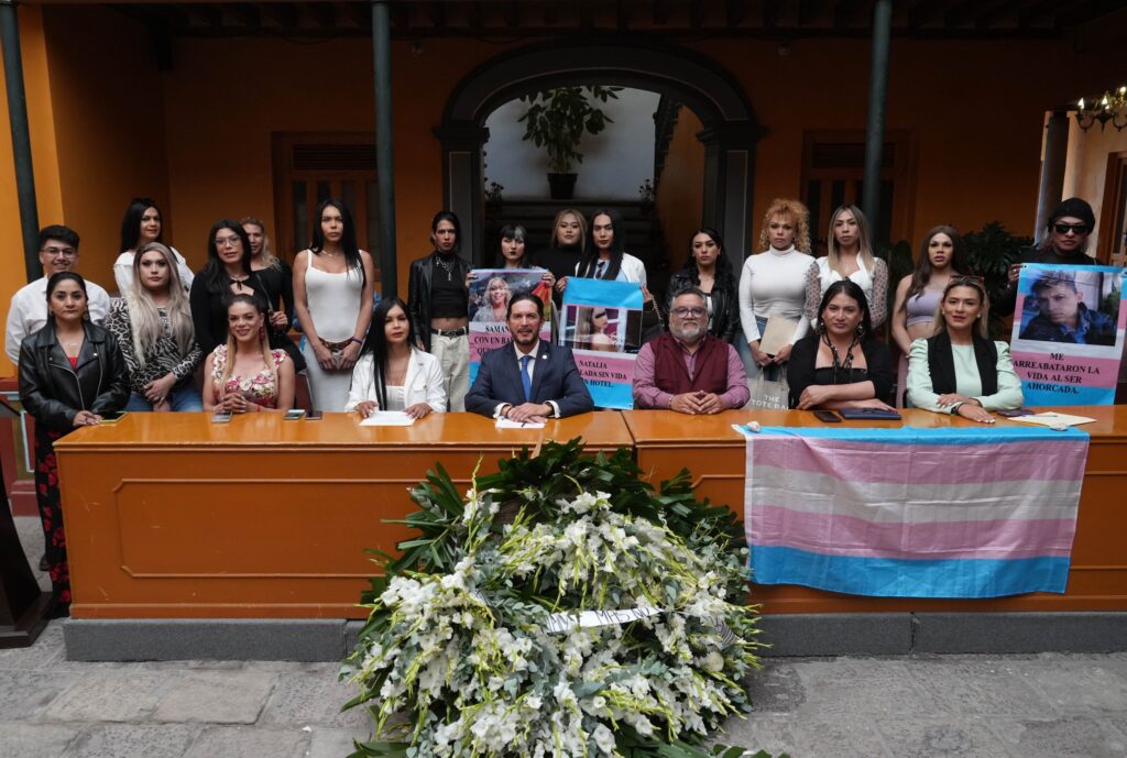 El presidente de la CODHEM Víctor Leopoldo Delgado Pérez encabezó una fotografía grupal durante el Foro/Conversatorio “TRANSgeneracional”, realizado en el marco del Día de la Remembranza TRANS, con participantes reunidos en torno a una mesa, una bandera trans visible y una ofrenda floral al frente, en un acto orientado a fortalecer la memoria histórica y la lucha social de las personas de la diversidad sexual y de género.