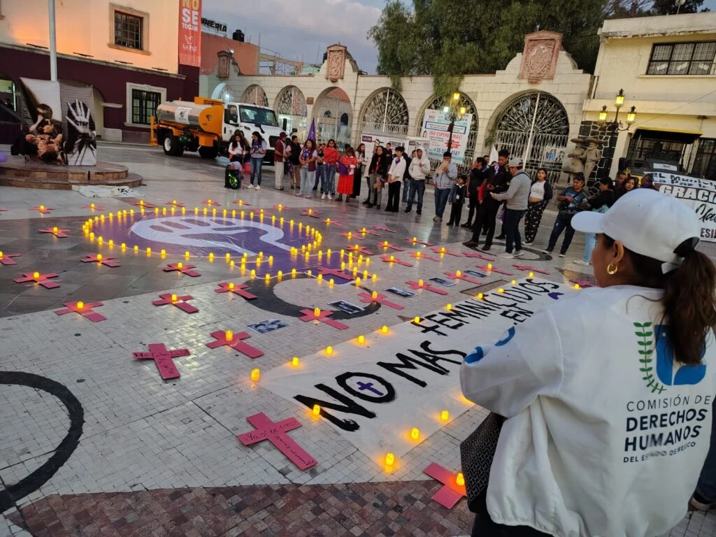 Persona con chamarra institucional de la Comisión de Derechos Humanos del Estado de México observa una concentración pública realizada en una plaza al atardecer. En el piso se ven veladoras encendidas, cruces rosas, carteles y una manta con el texto visible “No más feminicidios”, mientras un grupo de personas permanece alrededor del memorial.