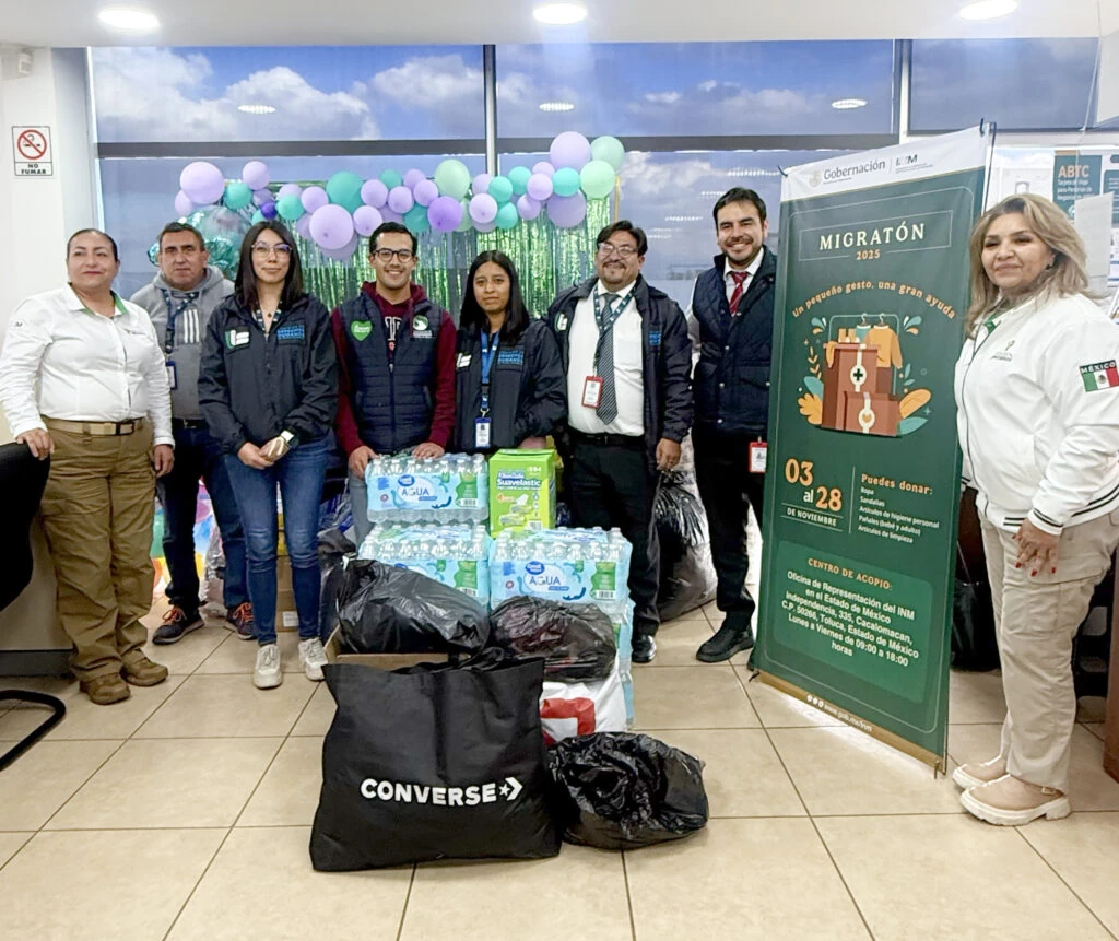 Grupo de personas posa en un centro de acopio junto a paquetes de agua, bolsas con donativos y otros artículos reunidos. Al fondo se observan globos decorativos en tonos verde y morado, así como un banner con el texto “Migratón 2025”, la frase “Un pequeño gesto, una gran ayuda” y fechas de recepción de donativos del 03 al 28 de noviembre.
