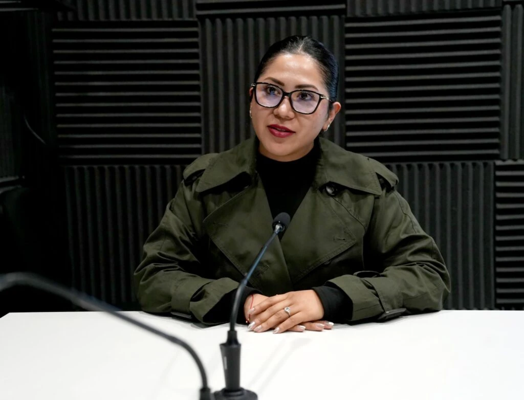 Persona sentada frente a una mesa de grabación o entrevista, con un micrófono colocado al centro. La persona porta gafas y una chaqueta verde oscura, mientras al fondo se observan paneles acústicos negros dispuestos en patrones geométricos. La imagen muestra un entorno acondicionado para la realización de programas, entrevistas, grabaciones de audio o actividades de comunicación institucional.