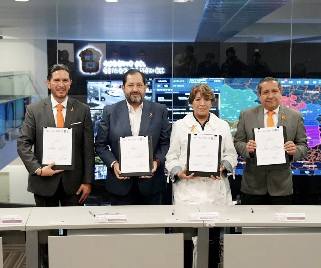 El presidente de la CODHEM Víctor Leopoldo Delgado Pérez consolida alianzas clave por los derechos humanos en un acto de firma de documentos institucionales; cuatro personas sostienen carpetas con documentos frente a pantallas de monitoreo y mapas digitales, en un espacio que parece corresponder a un centro de control o coordinación gubernamental.