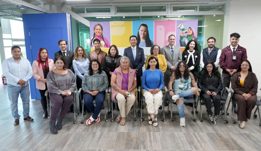 Personas participantes se reúnen alrededor de una mesa en forma de U durante una sesión de trabajo. Al frente de la sala se observa una pantalla con el título “Agenda LGBTTTIQA+”, así como banderas oficiales e integrantes de distintas instituciones y organizaciones dialogando sobre temas de inclusión y derechos humanos.