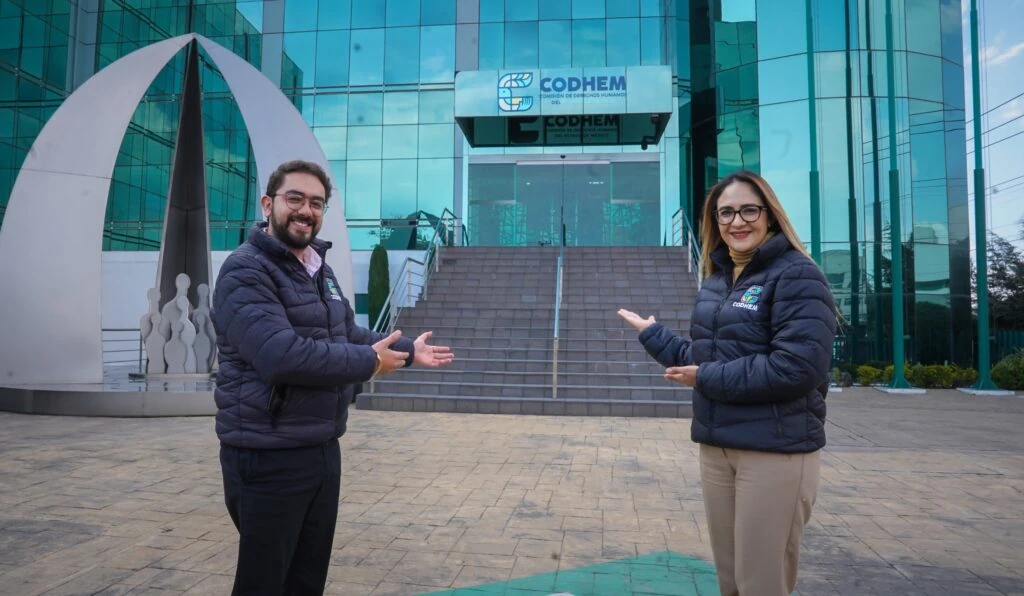 Dos personas con chamarras institucionales de la CODHEM señalan la entrada principal de un edificio con fachada de vidrio y logotipo visible. La imagen muestra el exterior de las instalaciones de la institución y contextualiza una actividad de atención u orientación ciudadana.