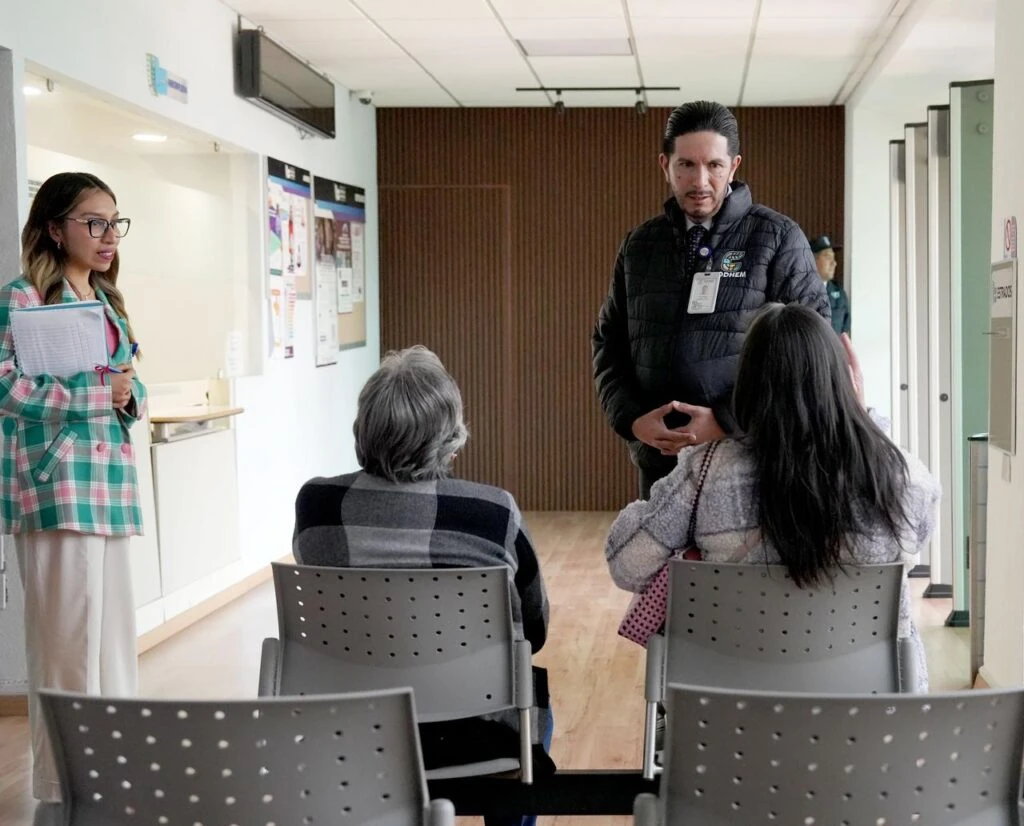El presidente de la CODHEM Víctor Leopoldo Delgado Pérez, Presidente de la Comisión de Derechos Humanos del Estado de México (CODHEM), ofrece orientación presencial a dos personas usuarias en una sala de espera. La imagen muestra una interacción directa en un espacio institucional iluminado, con sillas grises, ventanales, carteles informativos y una persona con libreta al lado izquierdo.
