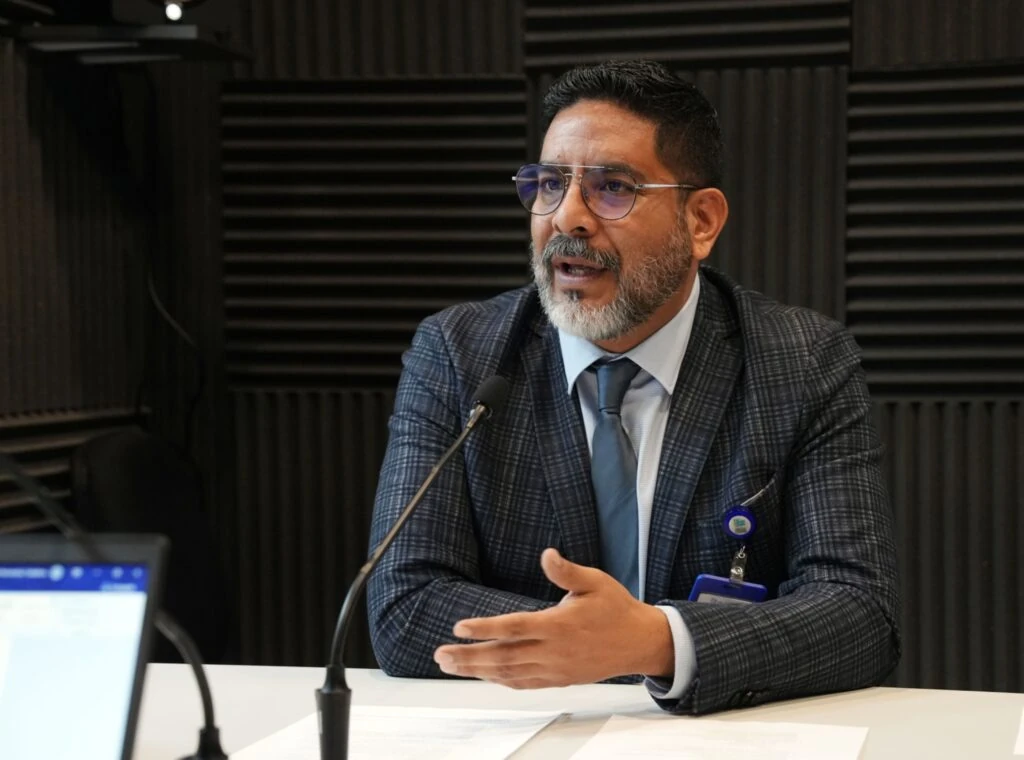 Persona con traje gris a cuadros, camisa clara, corbata azul y gafas habla frente a un micrófono durante una sesión de grabación o entrevista. La escena se desarrolla en un estudio con paneles acústicos negros en las paredes. Sobre la mesa se distinguen documentos impresos que sirven como material de apoyo, mientras en primer plano aparece parcialmente una pantalla de computadora.