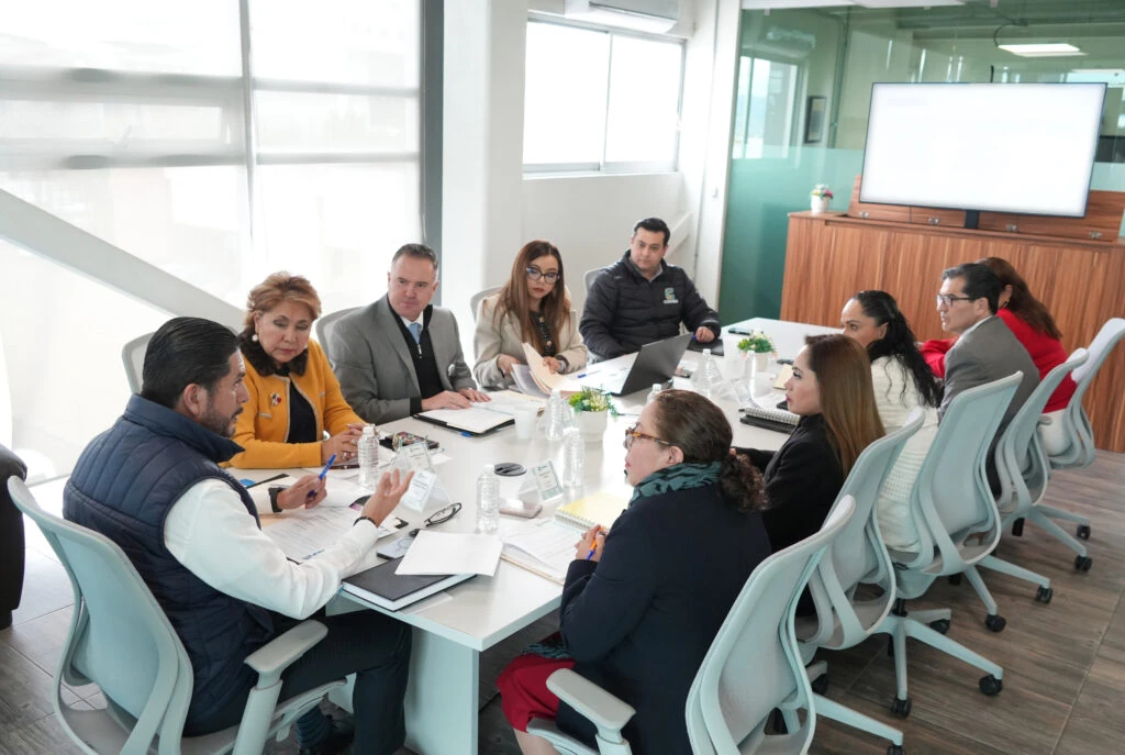 Personas participantes en una reunión de trabajo alrededor de una mesa blanca en una sala institucional, con documentos, libretas, botellas de agua, equipo de cómputo y una pantalla al fondo, en un espacio iluminado con ventanales y divisiones de cristal.