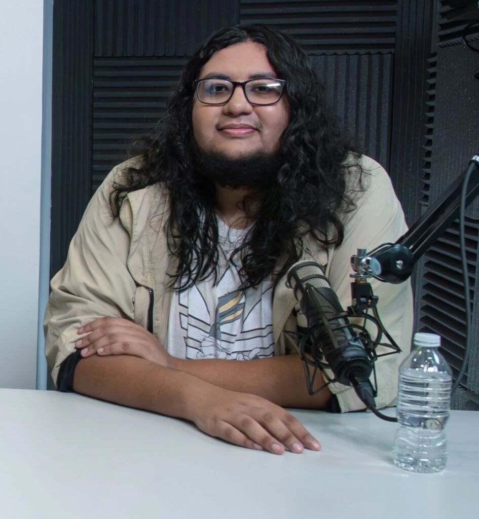 Persona sentada frente a una mesa en un estudio de grabación o podcast, con un micrófono montado en un brazo articulado y una botella de agua sobre la superficie. Al fondo se observan paneles acústicos negros en las paredes. La persona viste una chaqueta clara sobre una camiseta estampada y porta gafas.