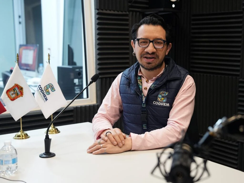 Persona participante en una grabación o entrevista dentro de un estudio de radio, sentada frente a un micrófono sobre una mesa. En el espacio se observan paneles acústicos y dos banderas de mesa, una con el logotipo de la CODHEM y otra con el escudo del Estado de México. La persona porta un chaleco institucional de la Comisión de Derechos Humanos del Estado de México.