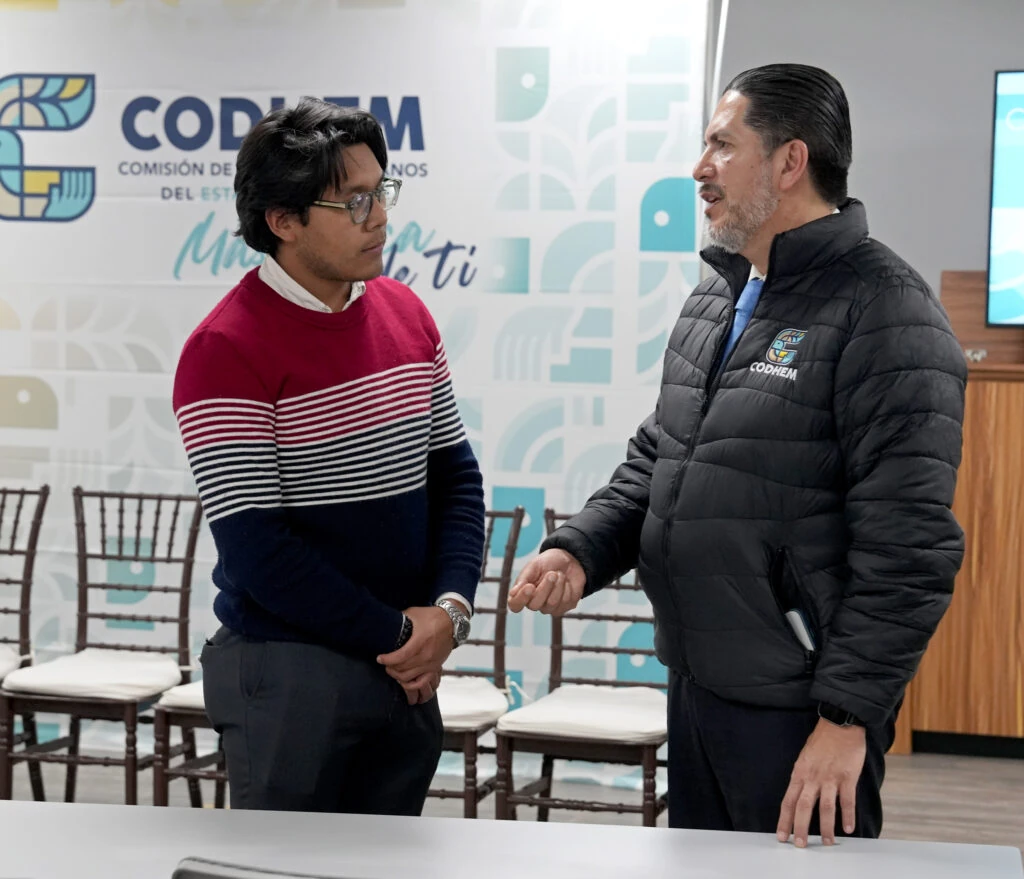 Integrante de la CODHEM conversa con un joven visitante en un salón institucional con el logotipo de la Comisión de Derechos Humanos del Estado de México al fondo. La escena forma parte de las visitas guiadas a la CODHEM, que acercan a las juventudes a aprender sobre la defensa de los derechos humanos.