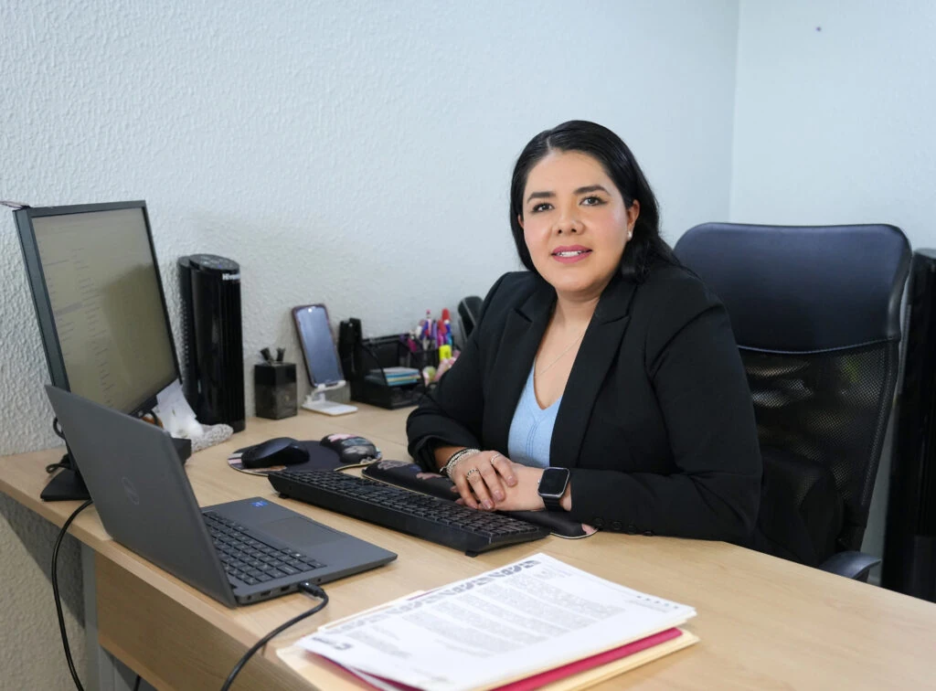 Persona sentada en un escritorio de oficina frente a un ordenador portátil, un monitor, teclado y documentos impresos; el espacio cuenta con paredes claras, silla de oficina y diversos accesorios de trabajo sobre la mesa.