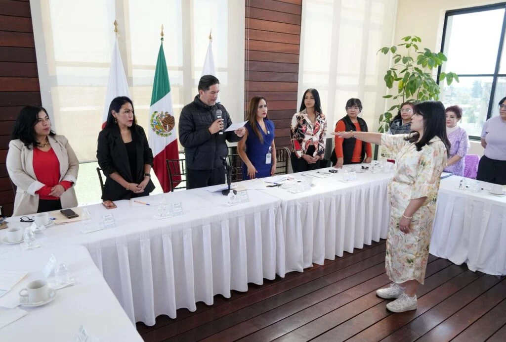Persona de pie con el brazo extendido durante una toma de protesta o acto protocolario en una sala de reuniones; alrededor se encuentran varias personas junto a mesas con manteles blancos, micrófono, documentos, tazas y banderas institucionales al fondo.