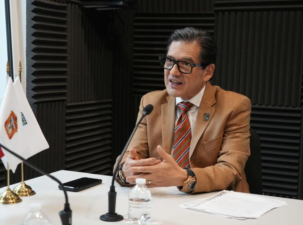 Persona sentada en una mesa durante una intervención con micrófono, con traje color café, camisa blanca, corbata a rayas y gafas; frente a la persona hay documentos, una botella de agua y dispositivos sobre la mesa, mientras al fondo se observan paneles acústicos negros y banderas institucionales.