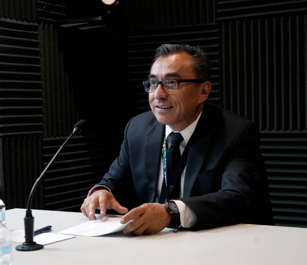 Persona servidora pública participa en una sesión institucional, sentada frente a un micrófono y documentos sobre una mesa, con traje oscuro, gafas y gafete; al fondo se observan paneles acústicos negros, en un contexto de diálogo relacionado con el fortalecimiento institucional y la protección de los derechos humanos.