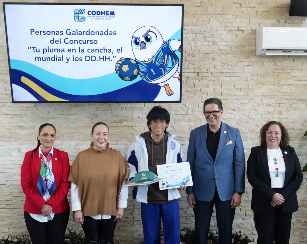 Fotografía grupal durante la premiación del concurso “Tu Pluma en la Cancha, el Mundial y los Derechos Humanos”, con personas representantes de la CODHEM y una persona galardonada que sostiene un reconocimiento y artículos alusivos al certamen. En la parte superior se observa una pantalla con el nombre del concurso y una mascota con balón de futbol, en el contexto de una ceremonia que destacó la participación de 207 estudiantes de secundaria y preparatoria de distintos municipios.