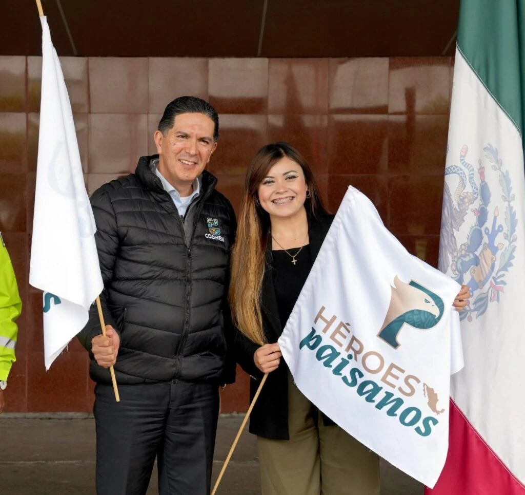 Dos personas posan con banderas del programa Héroes Paisanos y una Bandera de México durante una actividad institucional en el Aeropuerto Internacional de Toluca. La imagen acompaña el mensaje “Los derechos humanos no tienen fronteras: Víctor Delgado”, relacionado con acciones de protección, acompañamiento y respeto a los derechos humanos de personas en movilidad.