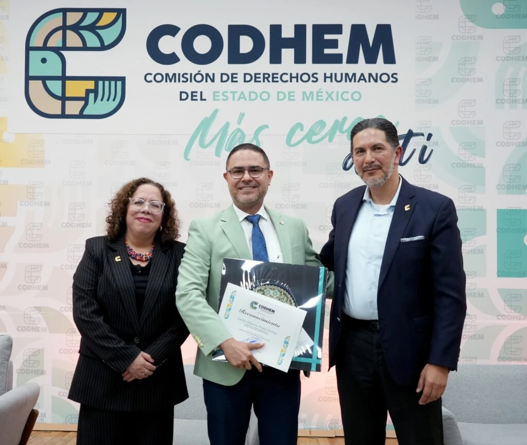 Participantes de una actividad de la CODHEM posan frente al emblema institucional durante la entrega de un reconocimiento. El evento destaca la relevancia de fortalecer la cultura de los derechos humanos mediante el conocimiento, la identificación y la defensa efectiva de estos en la vida cotidiana.