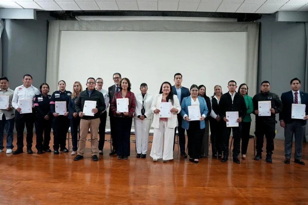Grupo de personas sobre un escenario durante una ceremonia de entrega de constancias. Varias personas sostienen documentos mientras posan frente a una pantalla de proyección en un salón de actos.