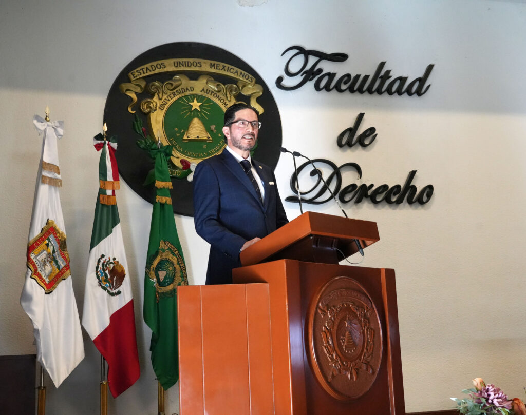 Persona realizando una intervención desde un podio en el auditorio de la Facultad de Derecho. Al fondo se observan el escudo universitario y las banderas de México y de la institución académica.