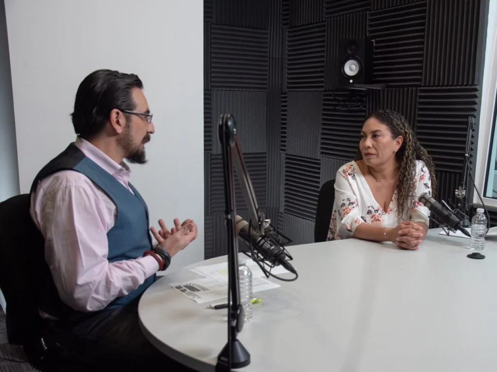 Dos personas participan en una conversación grabada en un estudio de pódcast. Ambas están sentadas frente a frente en una mesa blanca equipada con micrófonos montados en brazos articulados. El espacio cuenta con paneles acústicos oscuros en las paredes, hojas de trabajo sobre la mesa y botellas de agua junto a cada participante.