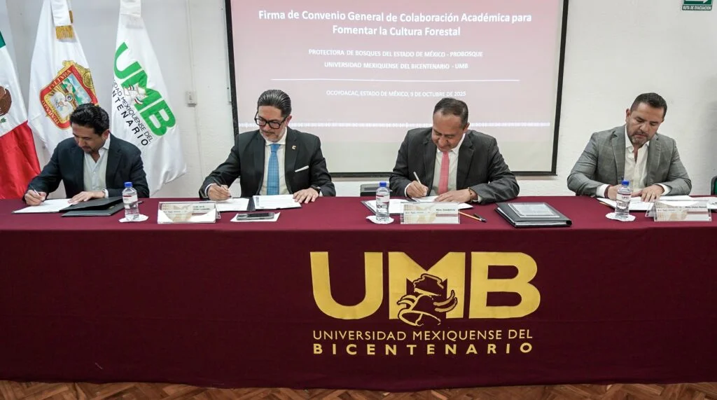 Cuatro representantes institucionales sentados detrás de una mesa con el logotipo de la Universidad Mexiquense del Bicentenario (UMB), mientras firman documentos durante la ceremonia de firma de un convenio general de colaboración académica para fomentar la cultura forestal. Al fondo se observa una pantalla con el título del evento y banderas institucionales.