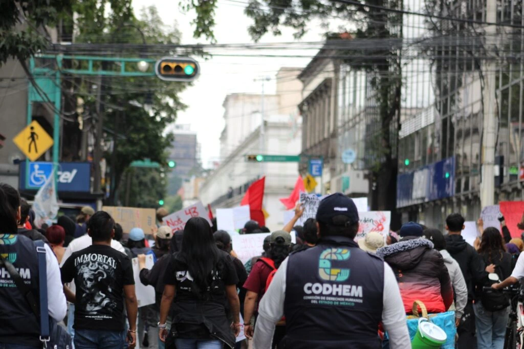 Personal de la CODHEM acompaña una movilización ciudadana que avanza por una avenida urbana. Las personas participantes portan pancartas, banderas y otros materiales de expresión pública.