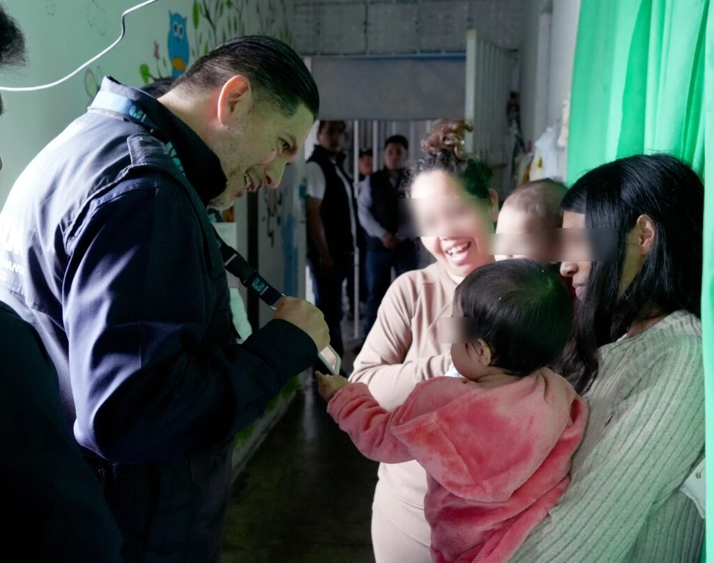 Integrante de CODHEM interactúa con un bebé sostenido por una persona adulta, mientras otras personas permanecen alrededor en un espacio interior.
