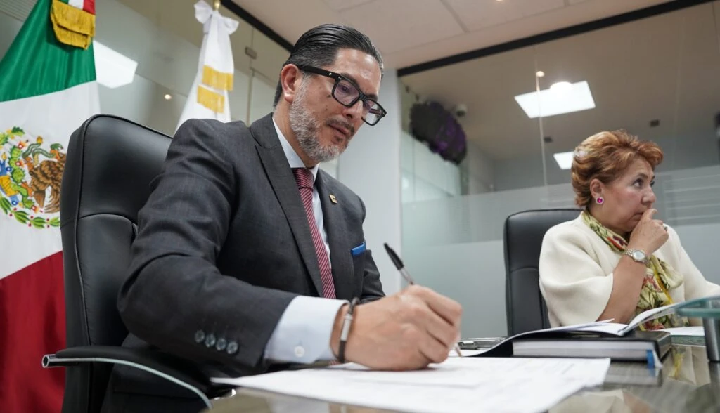 “Persona con traje oscuro y gafas escribe sobre un documento en una mesa de reunión. A un costado se encuentra otra persona sentada y, al fondo, se observan la bandera de México y una bandera institucional.”