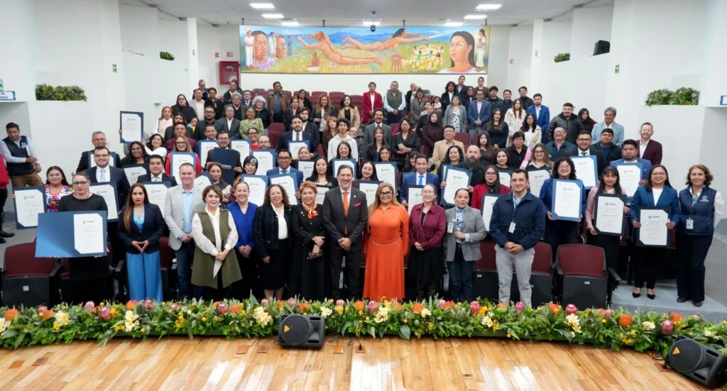 “Fotografía grupal en un auditorio institucional. Numerosas personas posan distribuidas entre las filas de asientos, varias sosteniendo reconocimientos enmarcados. Al fondo se observa un mural de gran formato y, en primer plano, un arreglo floral frente al escenario.”