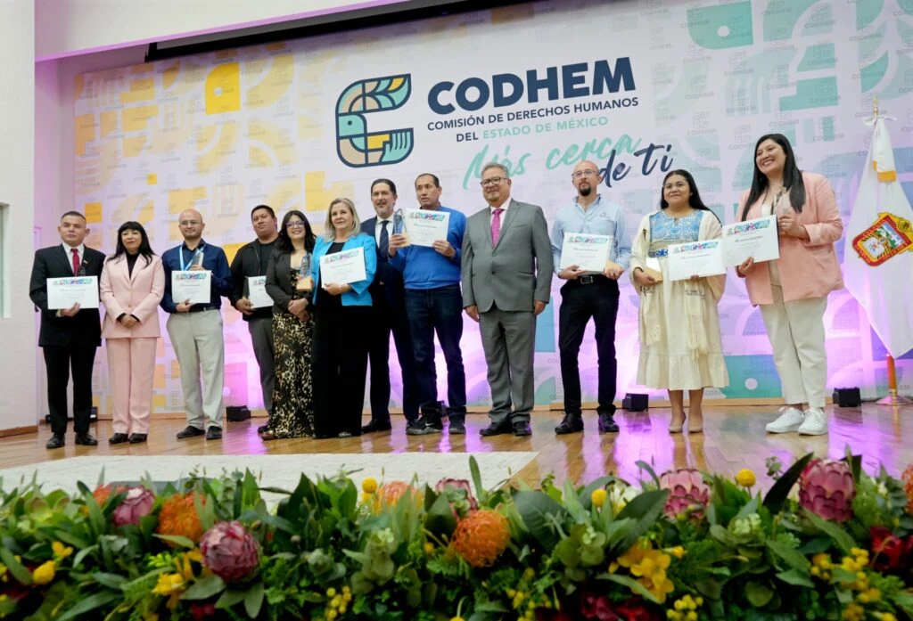 “Grupo de personas posa sobre un escenario durante una ceremonia de la CODHEM. Varias sostienen reconocimientos impresos y, al fondo, aparece la identidad gráfica de la Comisión de Derechos Humanos del Estado de México. Un arreglo floral ocupa la parte frontal del escenario.”