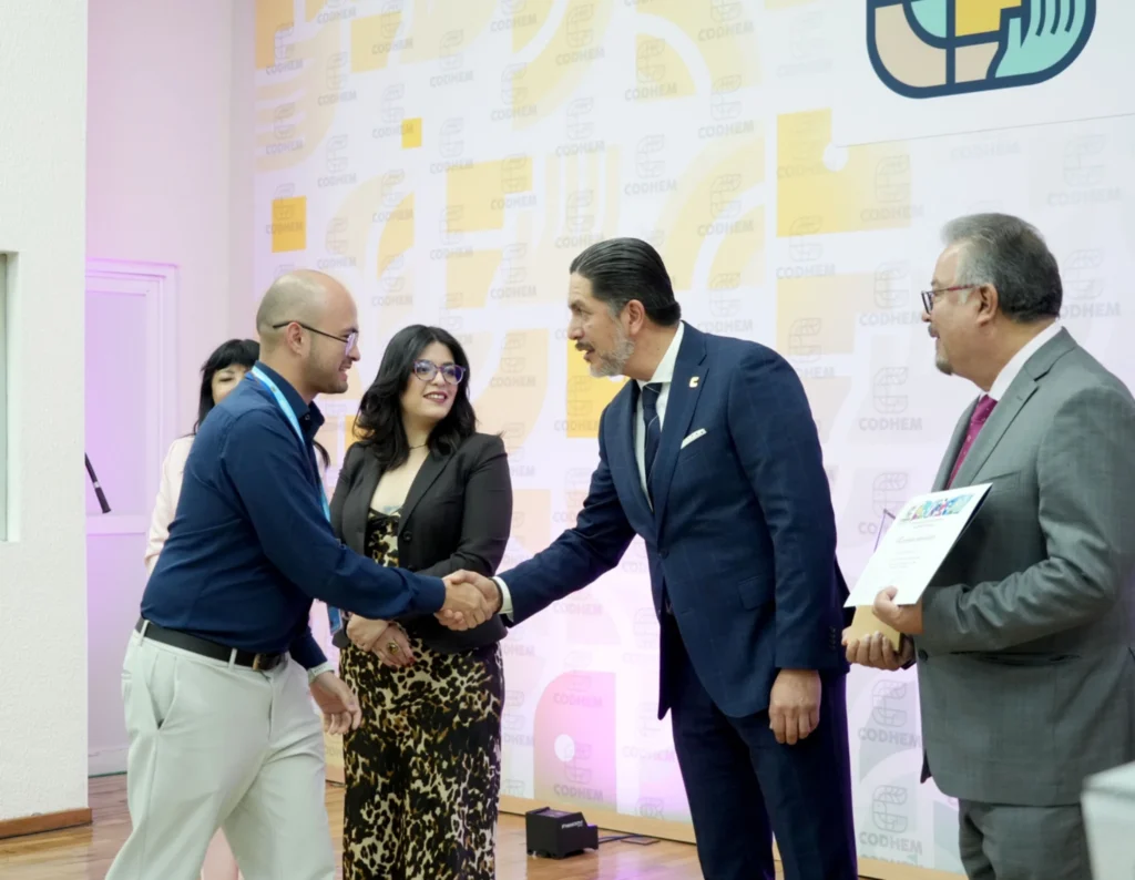 “Entrega de un reconocimiento durante un evento institucional de la CODHEM. Dos personas se estrechan la mano mientras otras observan; una de ellas sostiene un reconocimiento impreso. Al fondo se aprecia un muro con elementos gráficos de la CODHEM.”