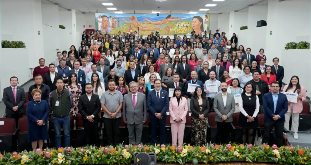 “Fotografía grupal en un auditorio durante un evento institucional. Un numeroso grupo de personas posa frente a las gradas; varias sostienen reconocimientos. En primer plano hay un amplio arreglo floral y al fondo se observa un mural.”