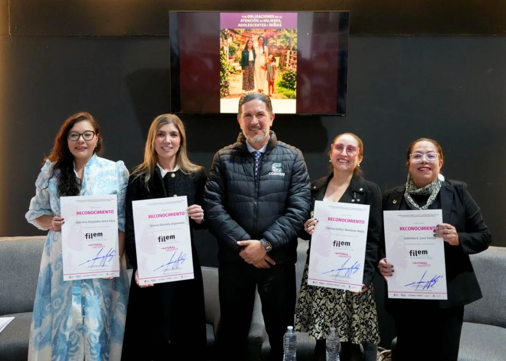 “Cinco personas posan durante una actividad institucional; cuatro sostienen reconocimientos de la FILEM. Al fondo, una pantalla muestra la portada de una publicación titulada ‘Tus obligaciones en la atención de mujeres, adolescentes y niñas’.”